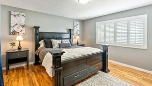 A bedroom with a dark wood bed, two bedside tables with lamps, a large window with shutters, and a floral painting on the wall.
