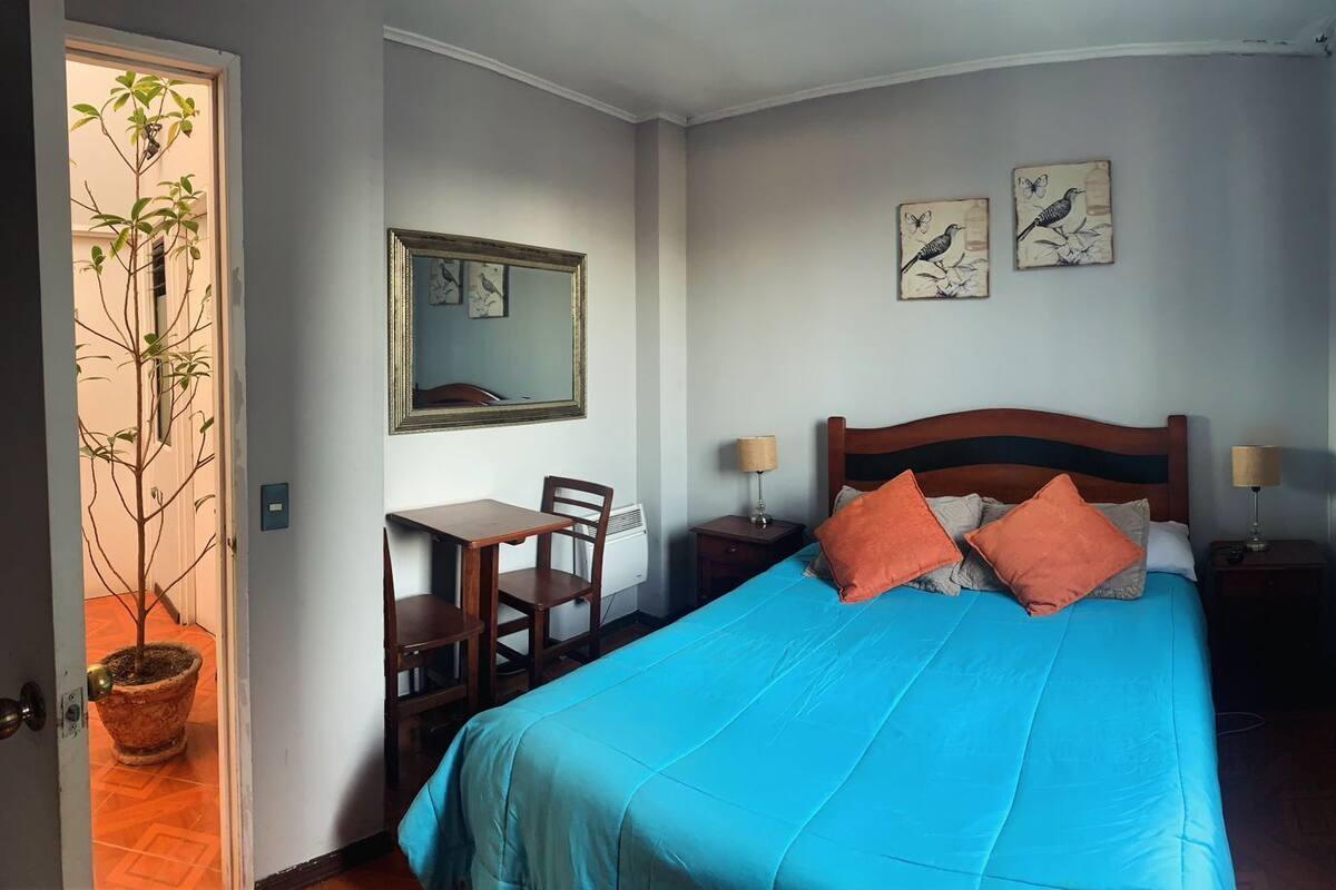 A bedroom with a bed, a desk, chairs, a mirror, and artwork on the wall.