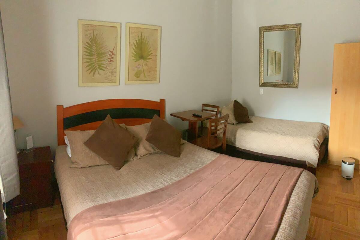 A room with two beds, a small table, chairs, a mirror, and two framed botanical illustrations on the wall.