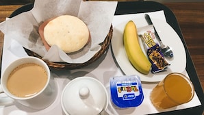 Una bandeja con una taza de café, un tazón de sopa, un plátano, una barra de chocolate y un recipiente de yogur.