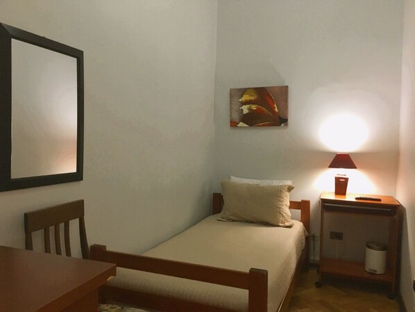 A small, neatly arranged bedroom with a single bed, a bedside table with a lamp, a chair, and a framed picture on the wall. - Residencial Universitaria (Santiago)