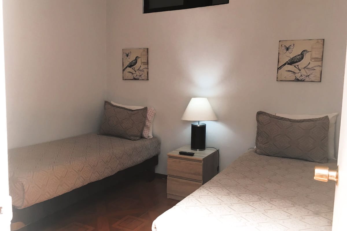 A small, simple room with two single beds, a nightstand with a lamp, and two wall-mounted pictures of birds.