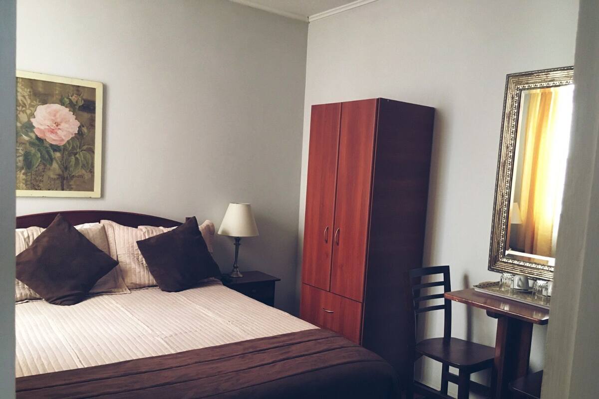 A bedroom with a bed, a wooden wardrobe, a desk with a mirror, and a lamp.