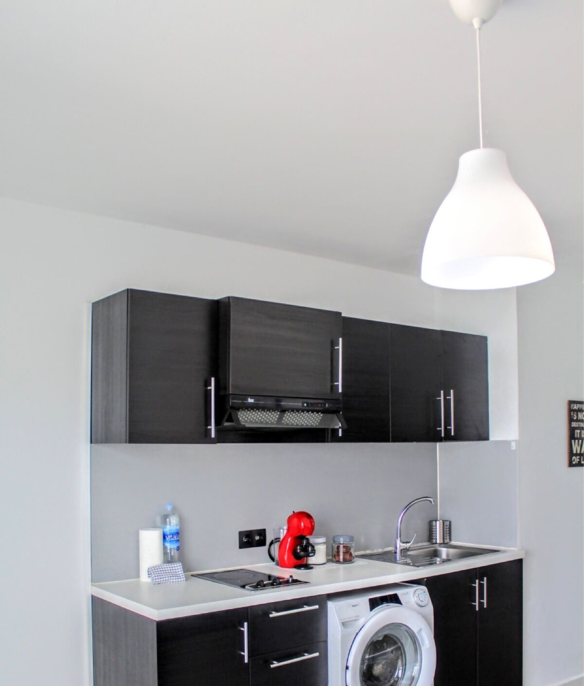 A compact kitchen with dark cabinets, a washing machine, a red coffee maker, and a white pendant light.