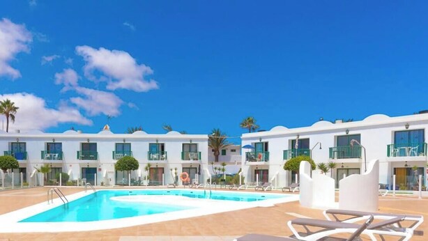 Zona de piscina con sillones de descanso, cielo azul despejado y un edificio con balcones.