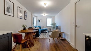 A modern living room with a sofa, coffee table, and chairs. There is a dining table with chairs, a lamp, and framed pictures on the wall. - Central 1BD Free Underground Parking (Cheltenham)