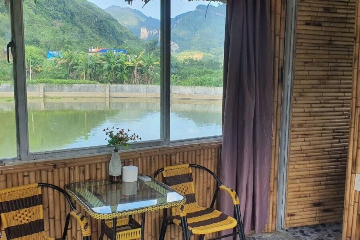 A room with a view of a lake and mountains, bamboo walls, a glass table with chairs, and a vase with flowers.