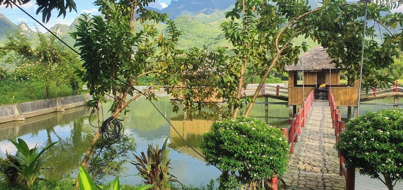 A wooden bridge leading to a thatched-roof structure over a calm water body, surrounded by lush greenery and mountains in the background.