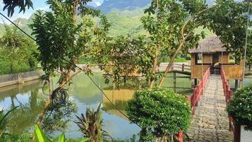 A wooden bridge leading to a thatched-roof structure over a calm water body, surrounded by lush greenery and mountains in the background.