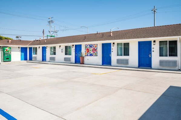A motel with multiple rooms, each with a blue door and a window. There is a green sign on the left side of the building.
