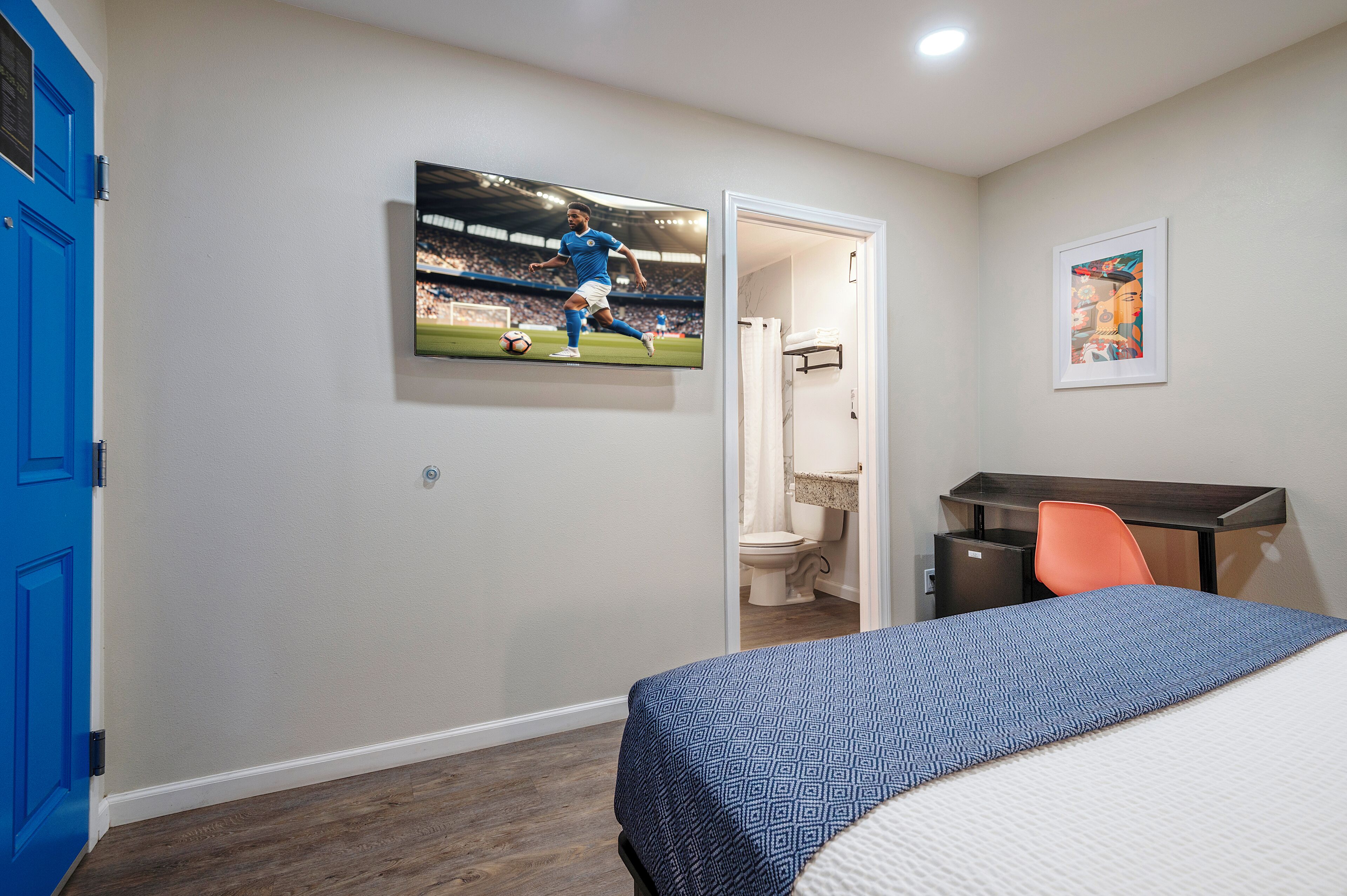 A hotel room with a bed, a desk, a chair, a television displaying a soccer match, and a framed artwork on the wall.