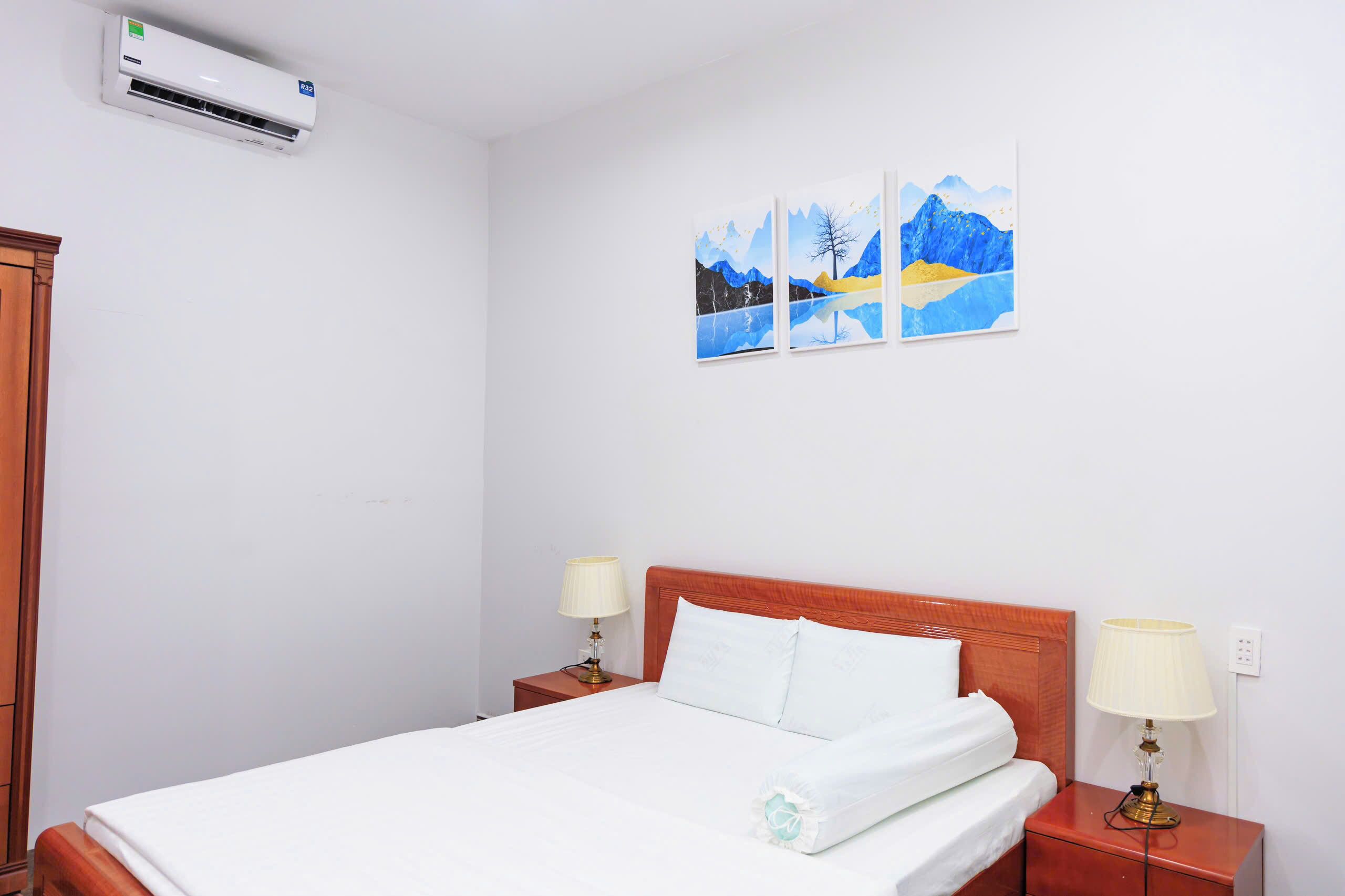 A hotel room with a bed, two bedside lamps, a wall-mounted air conditioner, and a painting above the bed.