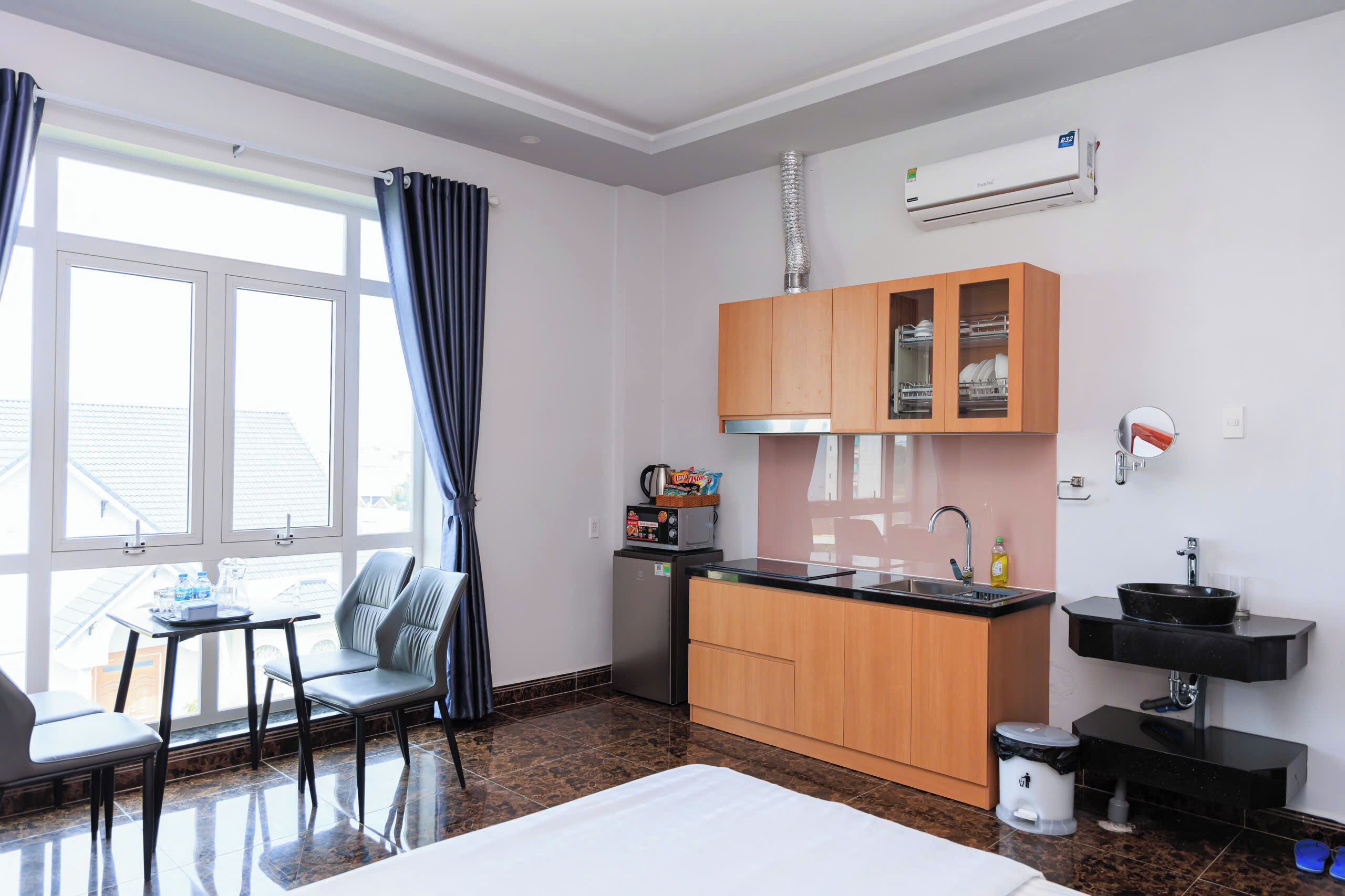 Ein kompaktes Hotelzimmer mit Küchenzeile, einem Essbereich mit Tisch und Stühlen, einem großen Fenster mit Vorhängen sowie einem Bett.