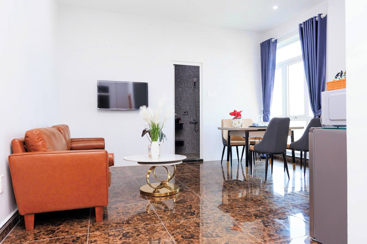 Un salon moderne avec un canapé en cuir, une table ronde dotée d’un pied doré, un téléviseur à écran plat fixé au mur, une petite plante en pot et un coin repas avec des chaises et une table.