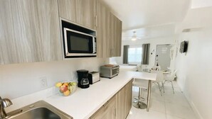 A modern kitchen with a built-in microwave, toaster, and coffee maker. There is a sink with a faucet, and a countertop with a fruit bowl. The kitchen leads to a dining area with a white table and chairs. - Studio | 2 Beds |  Kitchen | Rooftop| Near Beach (Miami beach)