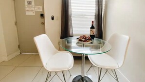 A small, modern room with a glass table and two white chairs, a bottle of wine, and a plate of food on the table.