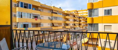 A balcony with a glass table and chairs, overlooking a yellow building with multiple balconies and air conditioning units.