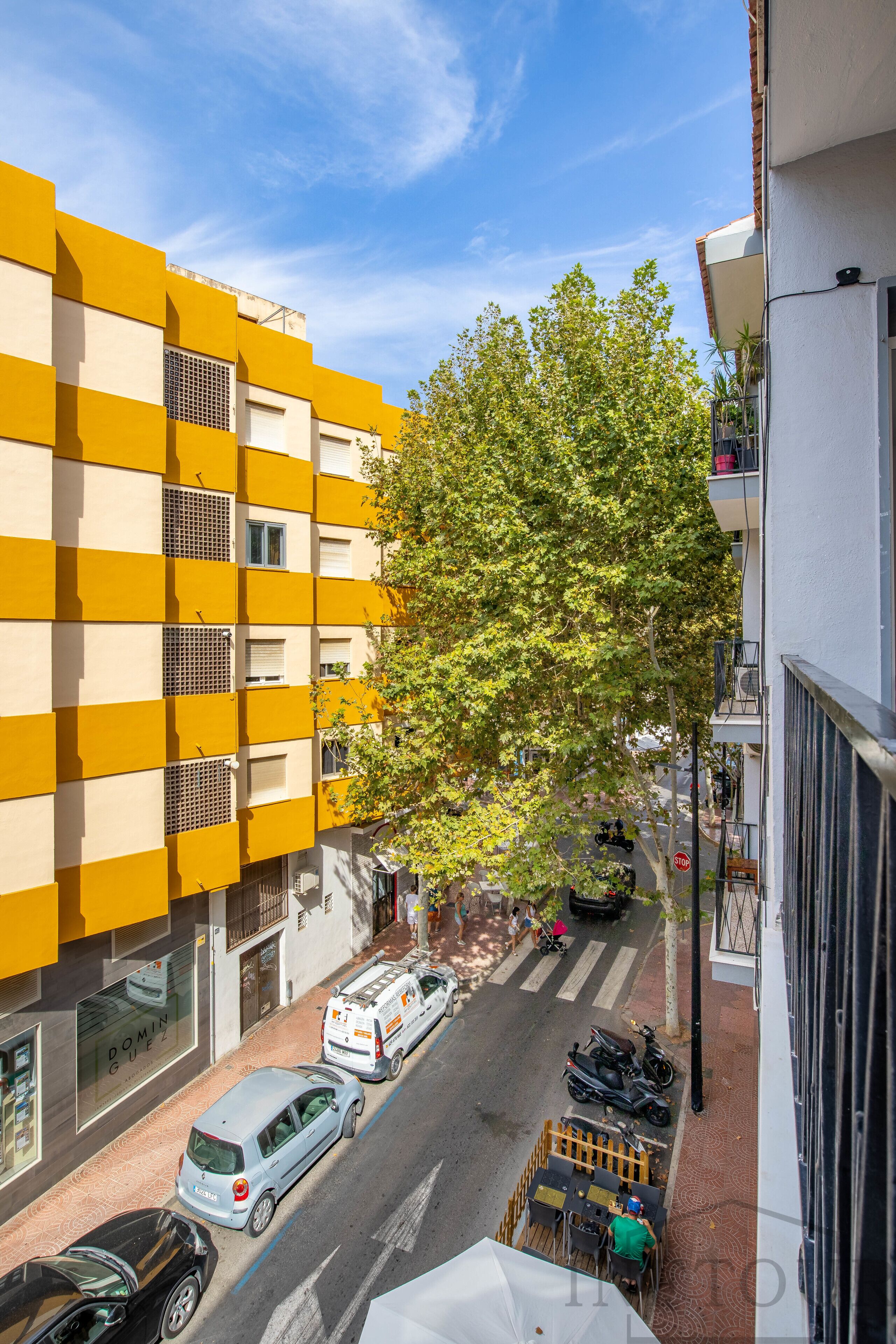 A street view with a yellow building, a tree, parked cars, and a café.