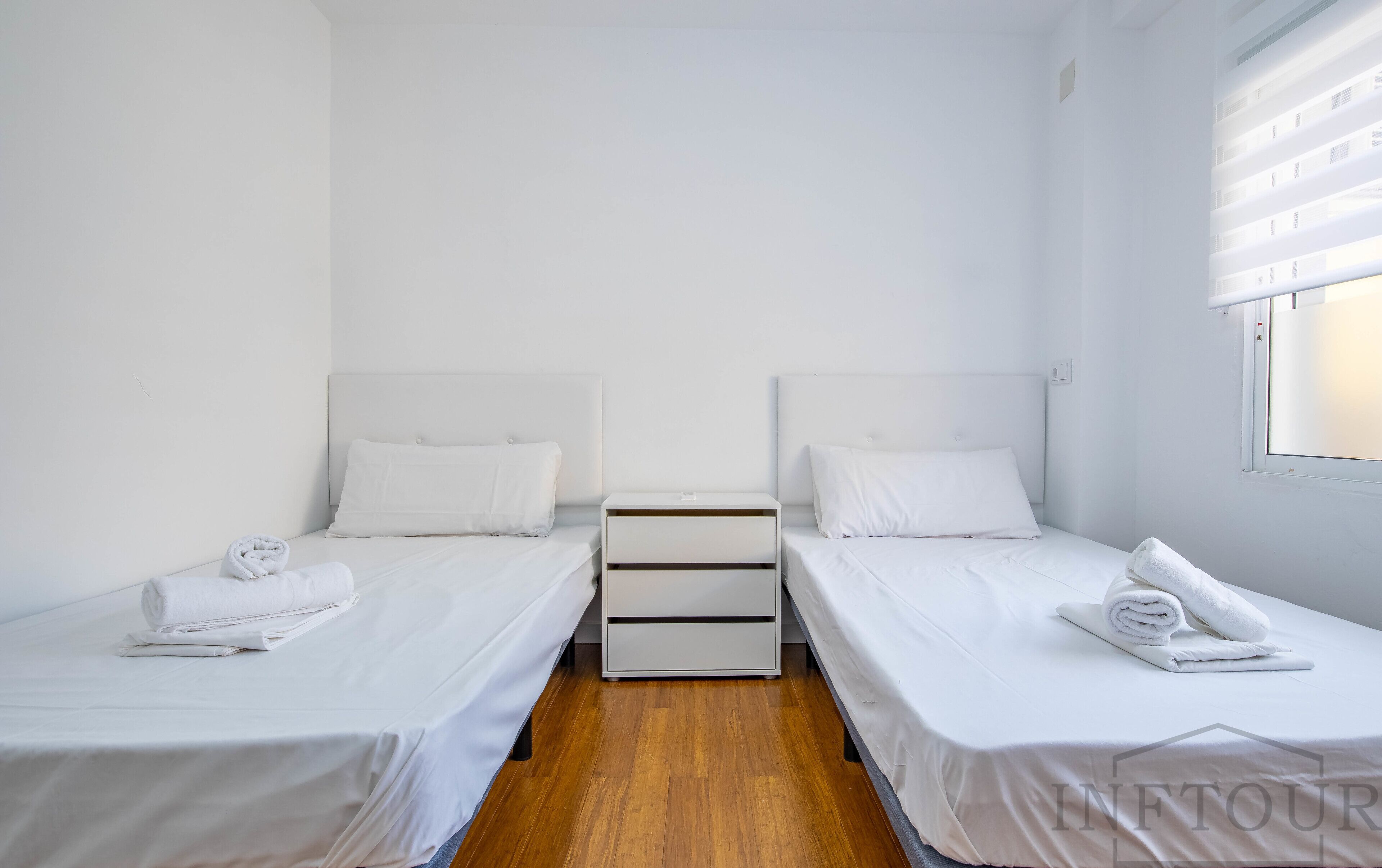 A clean, minimalist hotel room with two single beds, a small bedside table, and a window with blinds.