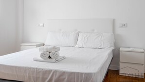 Ein ordentlich bezogenes Bett mit weißen Leinen und Kissen, ein kleiner Nachttisch mit Lampe und eine Wandsteckdose.
