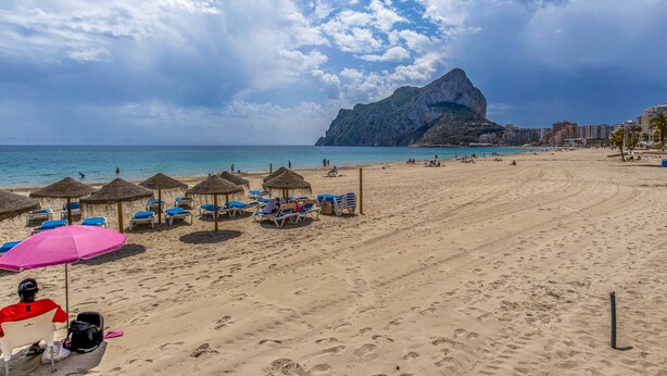 Ein Strand mit Sonnenschirmen, Liegestühlen und einem Berg im Hintergrund.