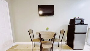 Un área de comedor con una mesa redonda y cuatro sillas, un televisor de pantalla plana en la pared, y un refrigerador a la derecha.