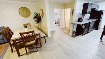 A modern kitchen with black cabinets and a white countertop, a dining area with a wooden table and chairs, and a hallway leading to another room.