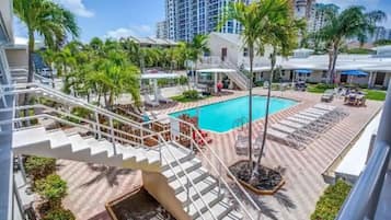 A hotel pool area with a staircase leading to the upper floors, surrounded by palm trees and lounge chairs.