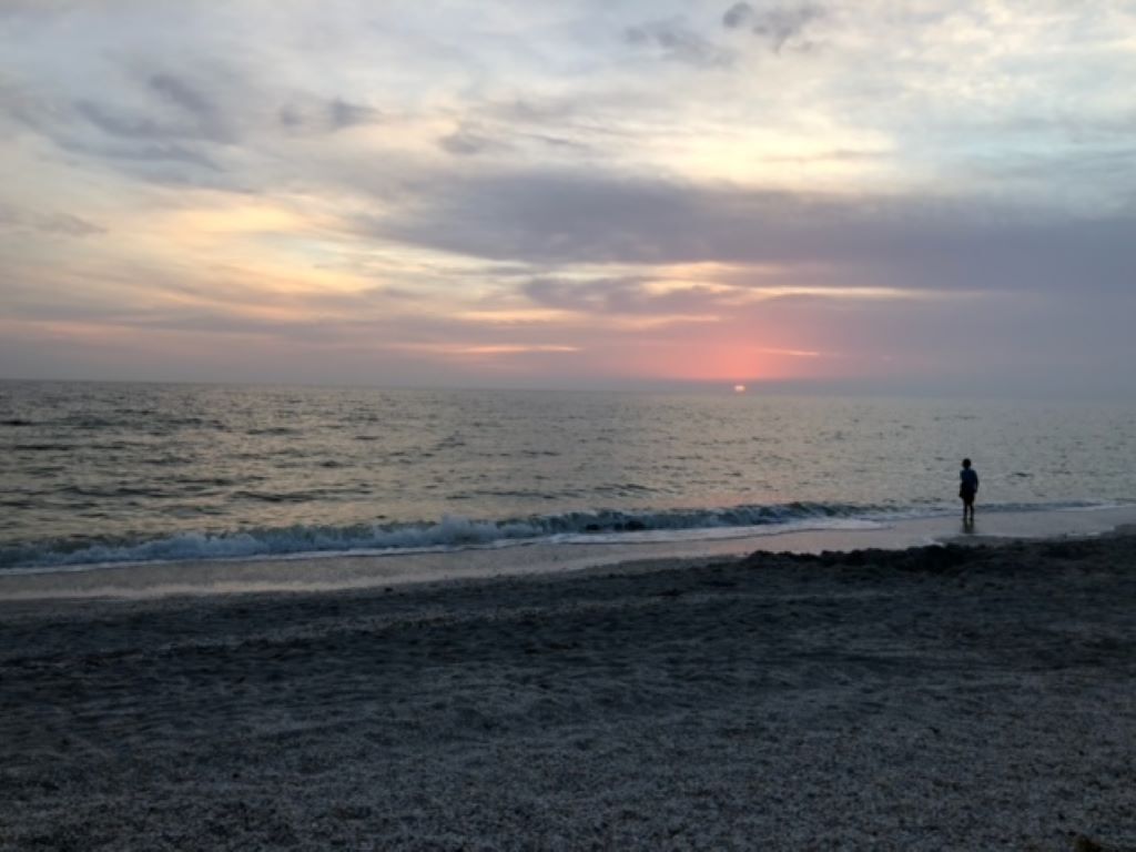 A beach with a person standing near the water's edge during sunset.