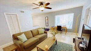A living room with a sofa, coffee table, and a ceiling fan. There is a window with blinds, a small table with a plant, and a TV stand. - Spacious 1 Bedroom Double Apt - Pool, Free Parking, Walk to Beach! (Lauderdale by the sea)