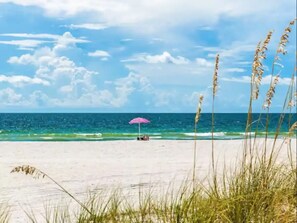 A beach with a clear sky, ocean waves, and a person under a pink umbrella.