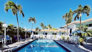 A swimming pool with lounge chairs and umbrellas, surrounded by palm trees and a building with a white fence. - Spacious 1 Bedroom Double Apt - Pool, Free Parking, Walk to Beach! (Lauderdale by the sea)
