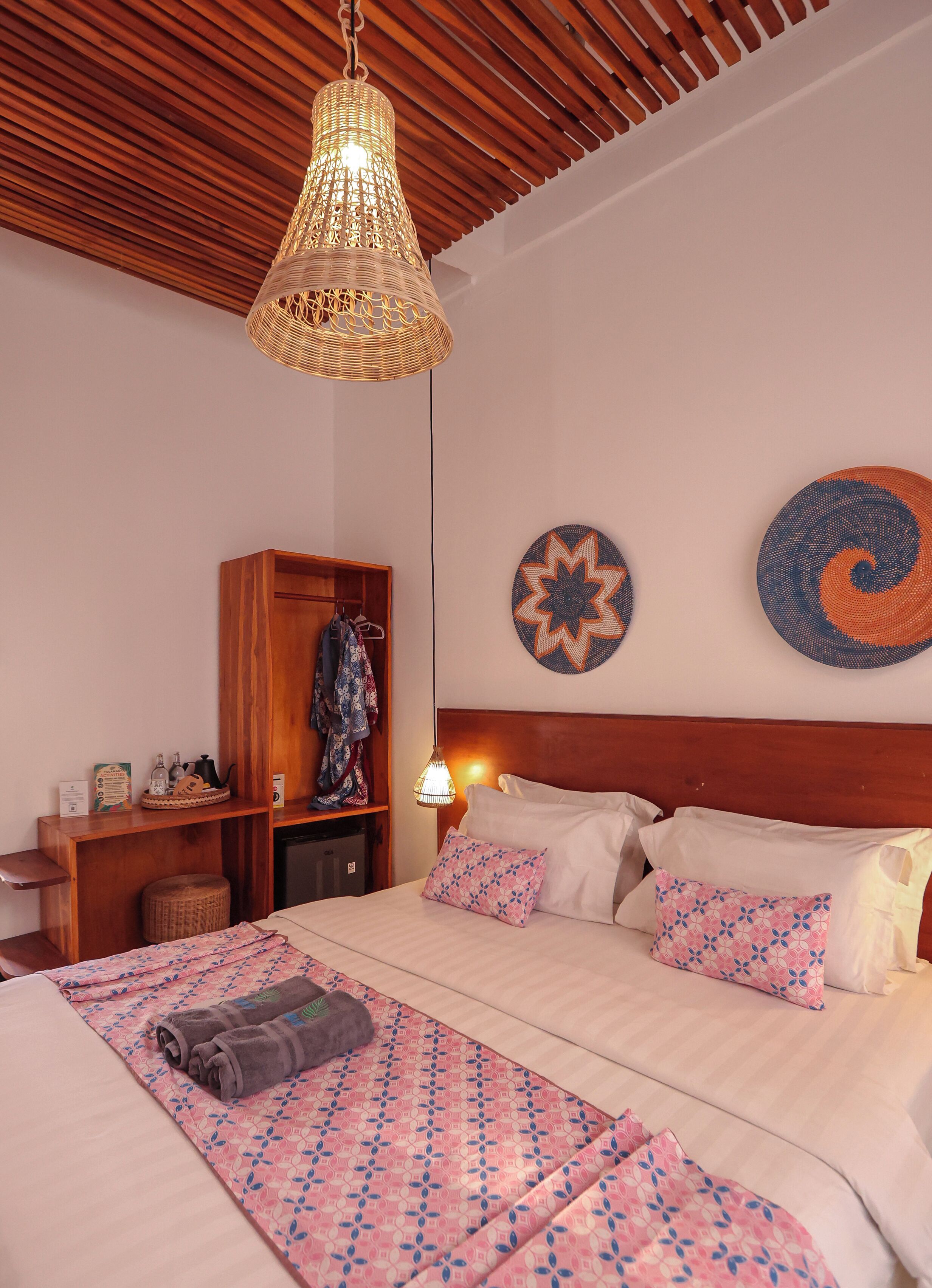 A bedroom with a bed, pillows, a hanging light fixture, a wooden headboard, a wardrobe, and a wall decoration.