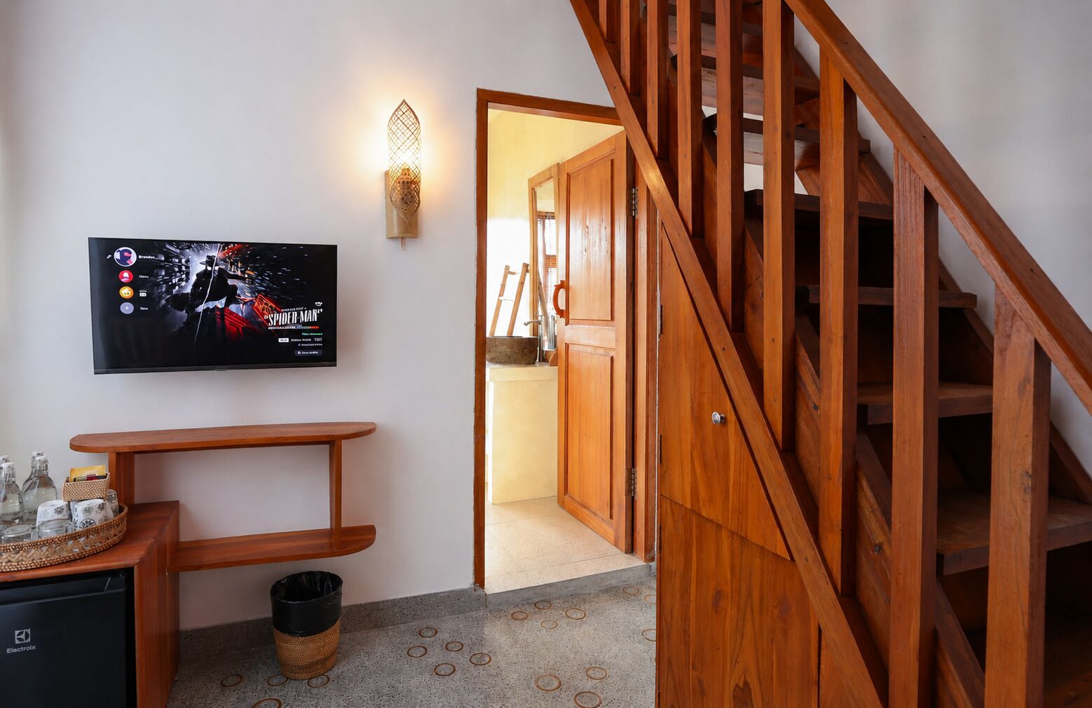 A staircase leading to a second floor, a wall-mounted television displaying a movie poster, and a wooden table with a basket and a trash bin.