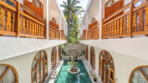 Un couloir d’hôtel avec une piscine centrale, des rampes en bois, des fenêtres en arc de cercle et un palmier.
