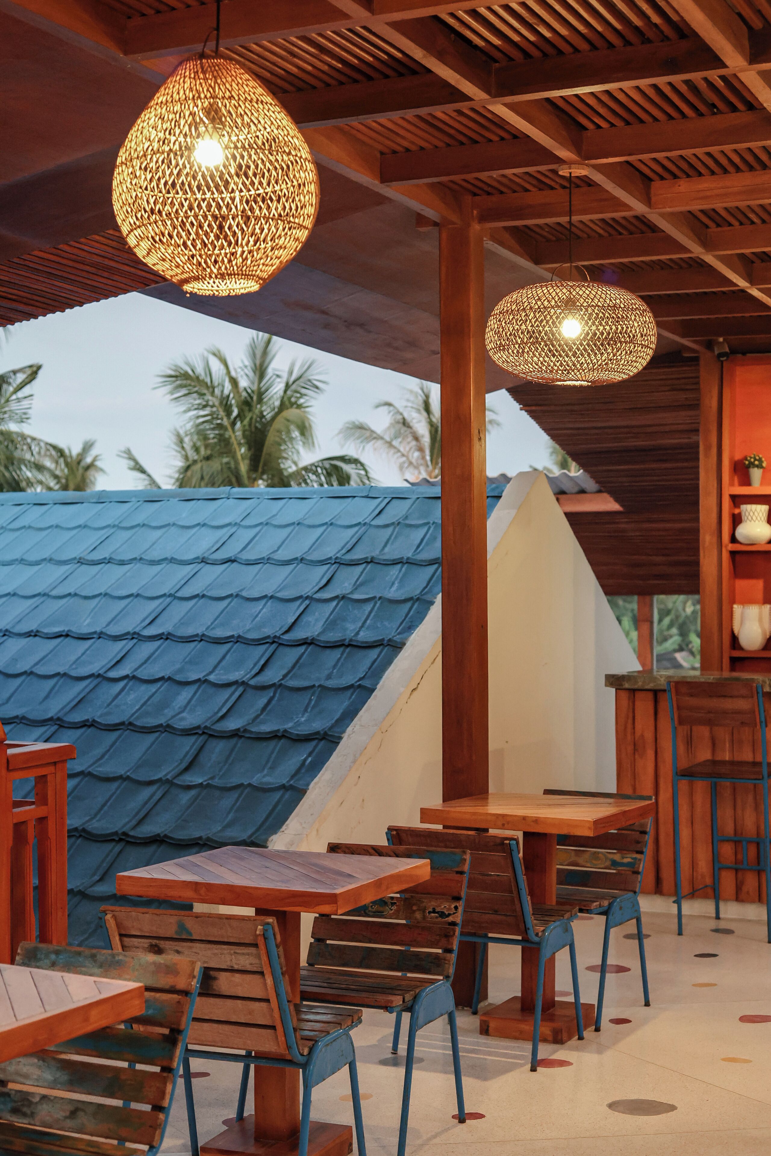 An outdoor seating area with wooden tables and chairs, a blue-roofed structure, and woven light fixtures.