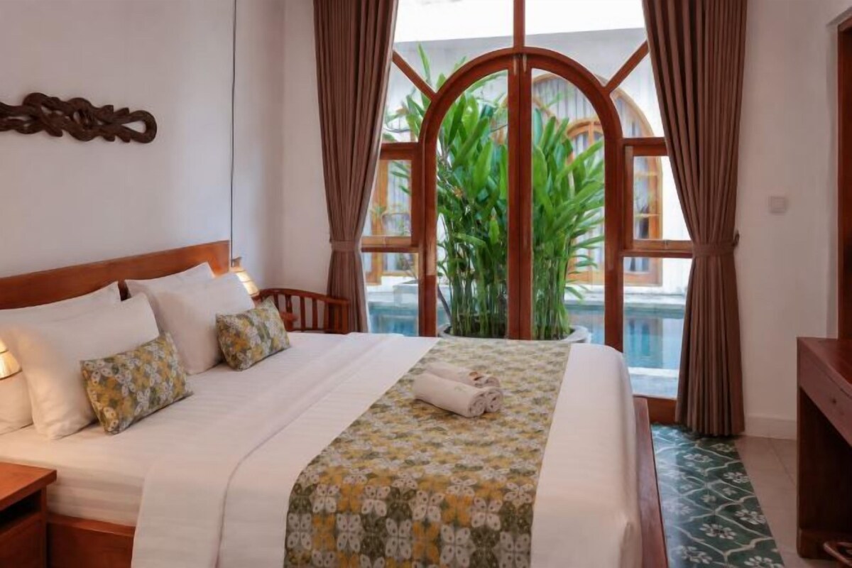 A hotel room with a large bed, a wooden headboard, a view of a pool, and a large window with curtains.