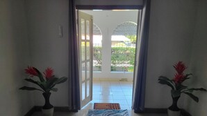A hallway with a tiled floor leading to a glass door. There are two potted plants with red flowers on either side of the door. A blue mat is placed on the floor in front of the door.