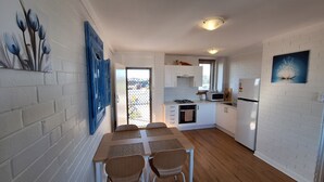 Une cuisine compacte avec un coin repas, des murs en briques blanches et une vue sur l’extérieur par la porte.