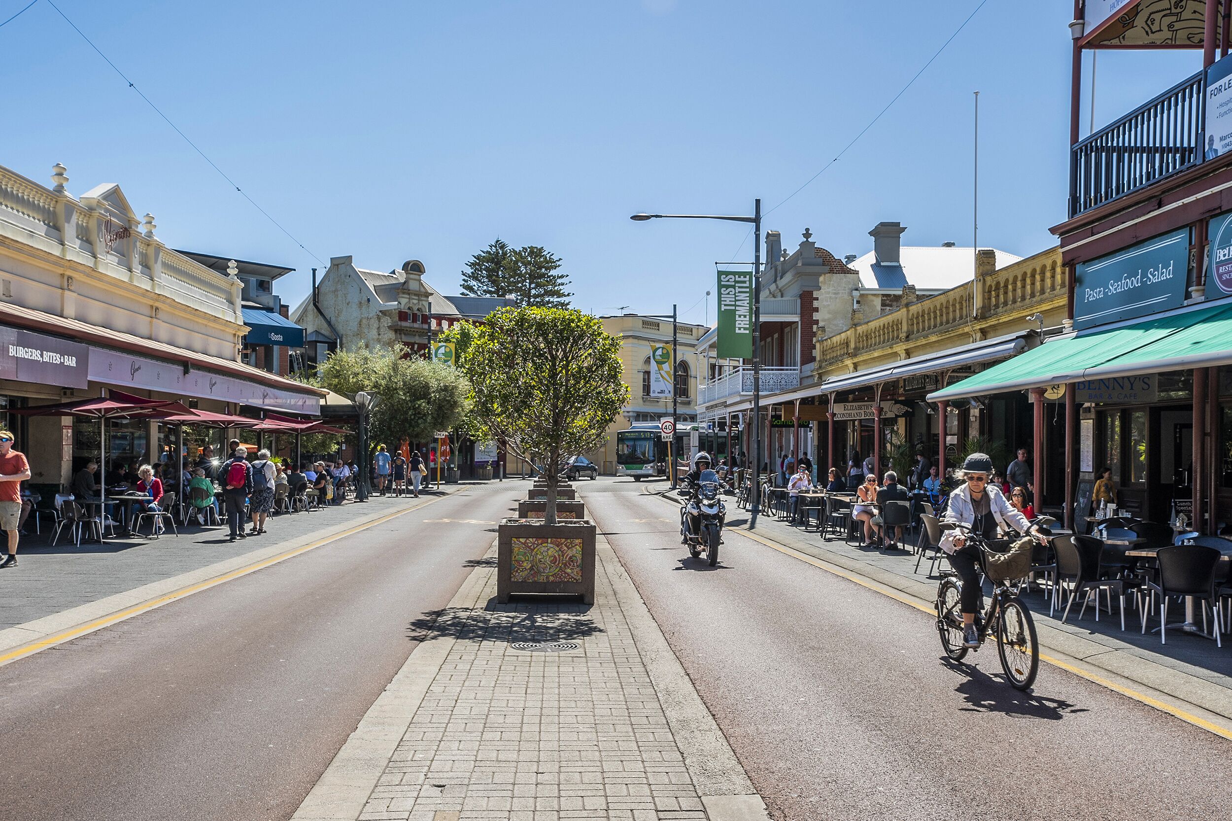 Rua com cafés e restaurantes, um ciclista na estrada, e um céu azul límpido.