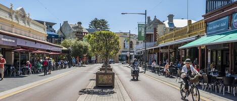 Rua com cafés e restaurantes, um ciclista na estrada, e um céu azul límpido.