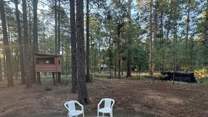 Ein Campingplatz mit einer runden Feuerstelle, umgeben von weißen Plastikstühlen, einer Holzbank und einer kleinen Struktur im Hintergrund.