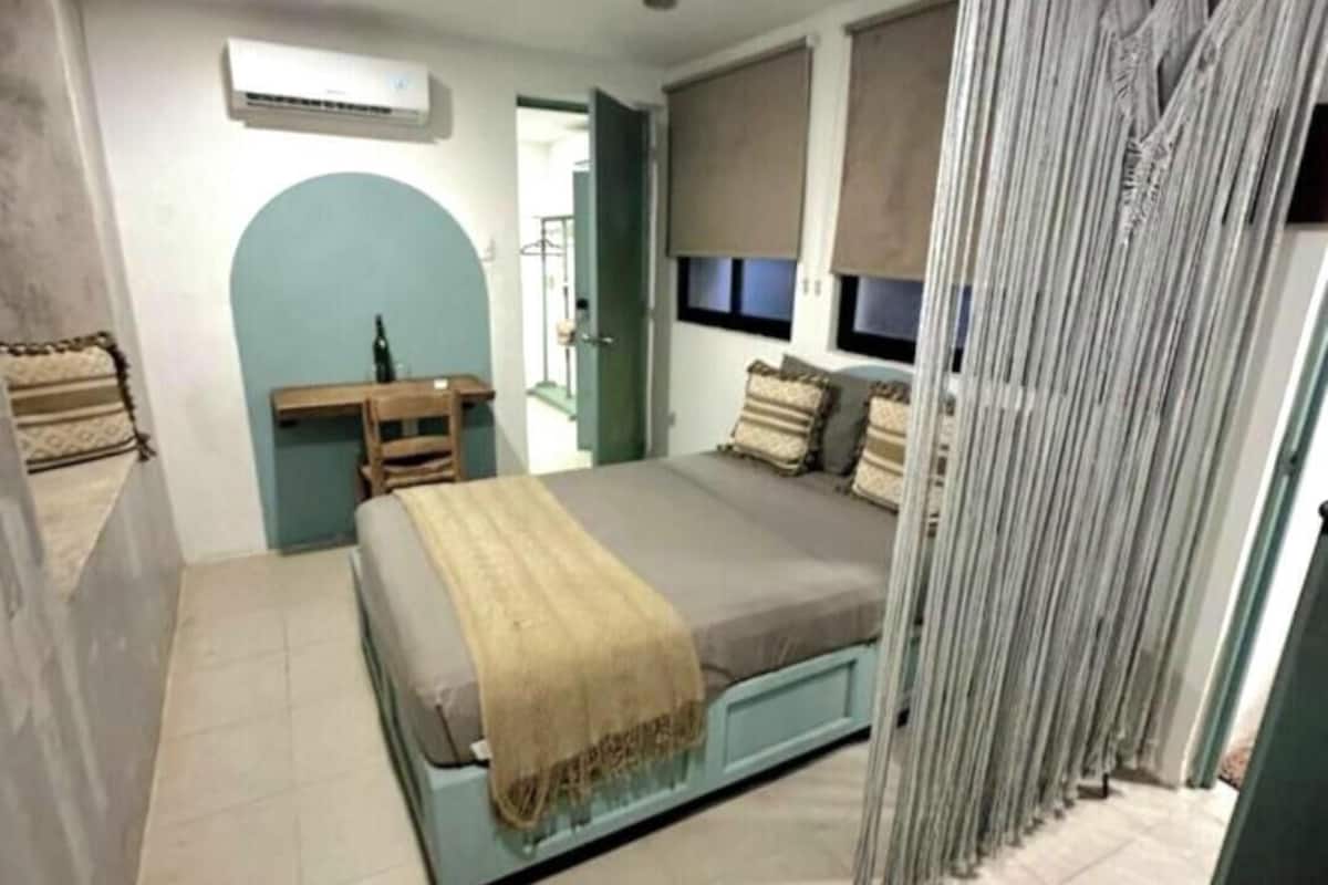 Un dormitorio con una cama, un escritorio y una silla. Se ve una ventana con persianas y cortina.