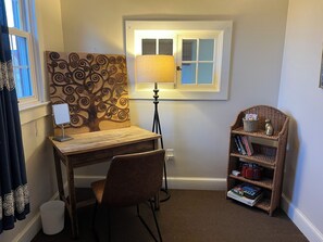 Ein Eckzimmer mit Schreibtisch, Stuhl, Lampe, Bücherregal und Fenster.
