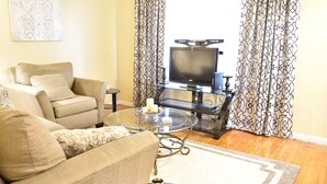 A living room with a beige sofa, a glass coffee table, a flat-screen TV on a stand, patterned curtains, and a wooden floor. - Cute 3br Ranch, Home Away From Home, Sleeps 6 (Detroit)