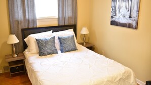 A neatly made bed with white and blue patterned pillows, a wooden nightstand with a lamp, and a painting on the wall. - Cute 3br Ranch, Home Away From Home, Sleeps 6 (Detroit)