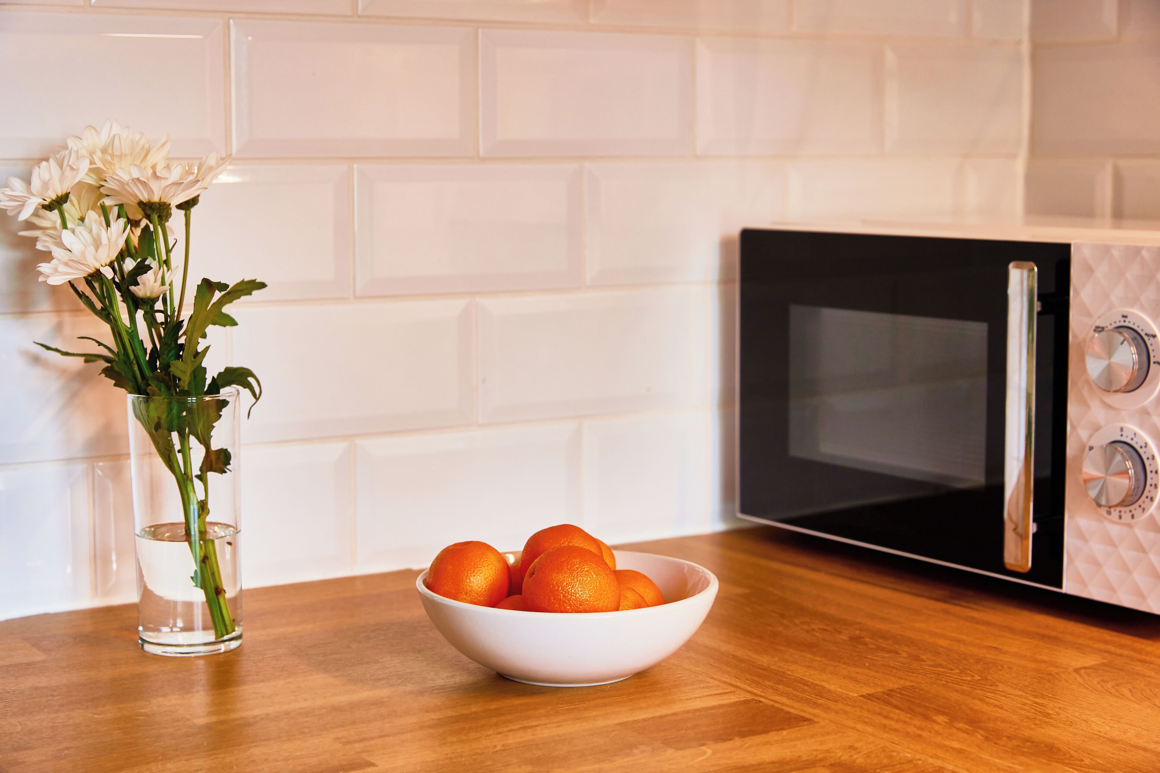Een keuken met een witte betegelde achterwand, een magnetron, een schaal oranjes en een vaas met bloemen.