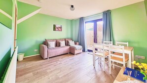 Ein Wohnzimmer mit einer Couch, einem Esstisch mit Stühlen, einem Bild an der Wand und einem Fenster mit Vorhängen.