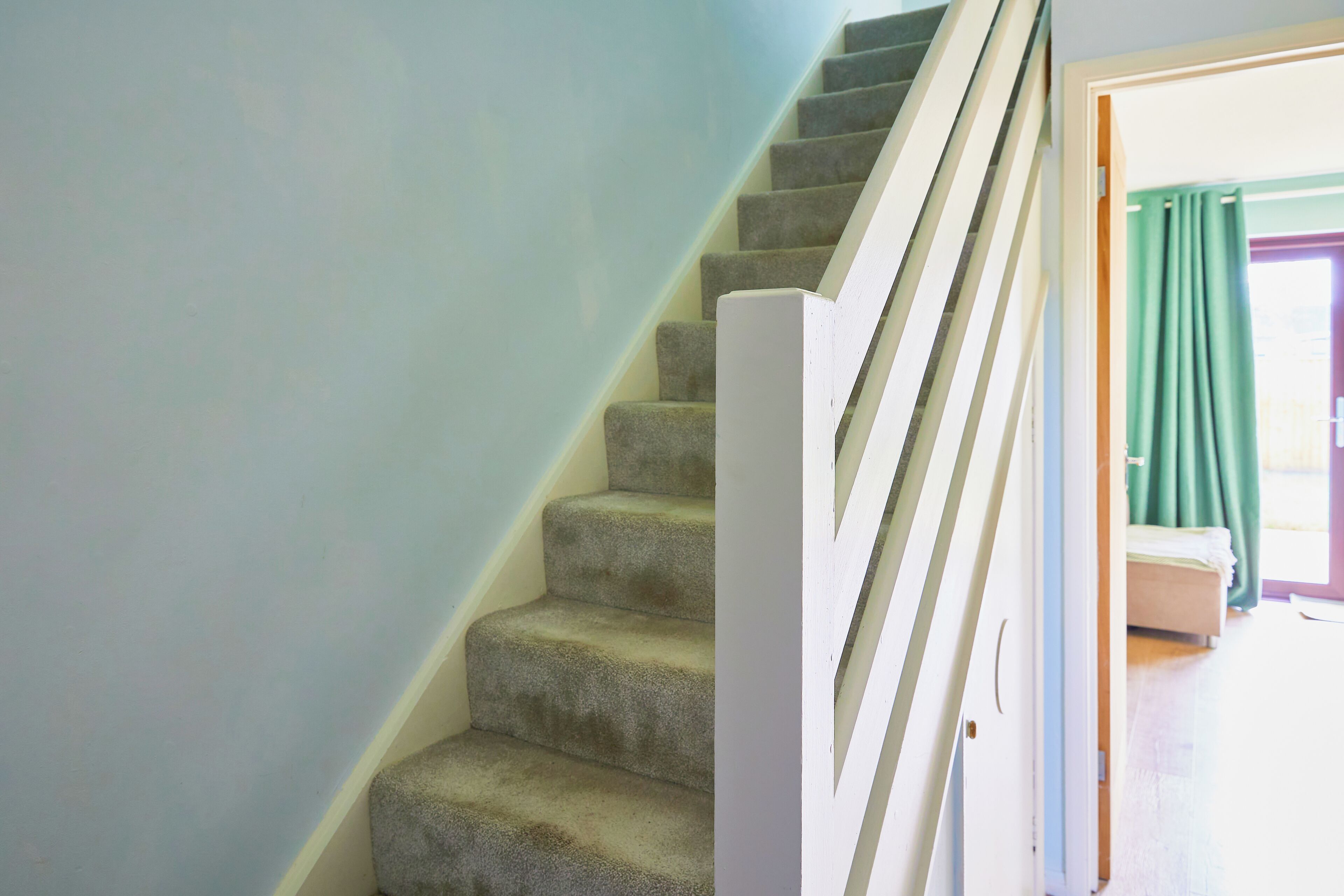 Un escalier recouvert de moquette, avec une rampe blanche, mène à une pièce où se trouvent un lit et une fenêtre ornée de rideaux verts.
