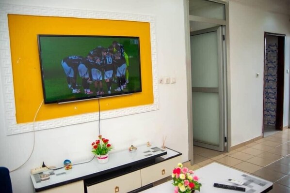 A living room with a flat-screen TV mounted on a yellow wall, a white console table with decorative items, and a sliding glass door leading to another room.